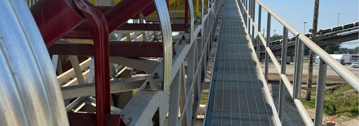 Galvanized CHCS Series ¾ corrugated conveyor covers with the open side facing a grated walkway, exposing red idler frames and yellow idlers for maintenance access