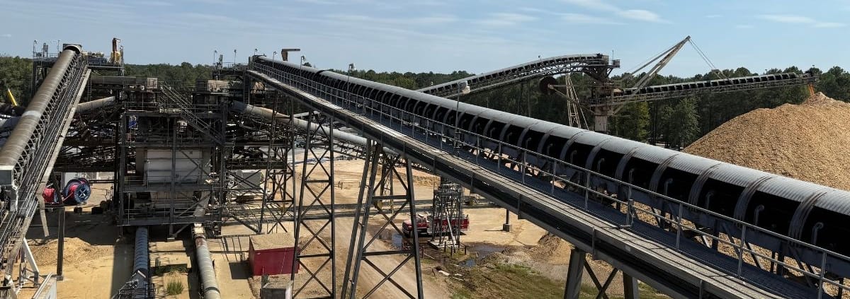 Long inclined conveyor fitted with CHCS ¾ corrugated covers at an outdoor bulk material site, open side revealing the belt, with additional covered conveyor lines and a material pile in the backgroundLong inclined conveyor fitted with CHCS ¾ corrugated covers at an outdoor bulk material site, open side revealing the belt, with additional covered conveyor lines and a material pile in the background
