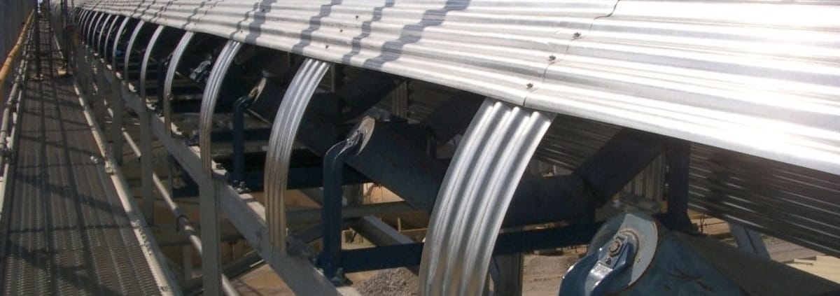 Galvanized CHLC Series conveyor covers with longitudinal corrugations running parallel to the belt, open side facing a grated walkway with blue idler frames and belt visible