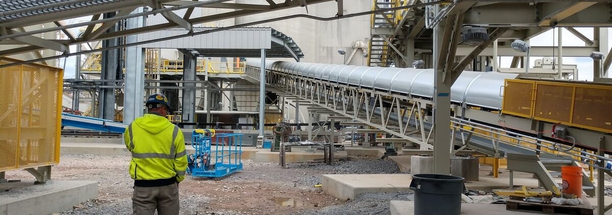 Crew member in high-visibility gear at an industrial site during conveyor cover installation, with a covered conveyor run and lift equipment behind him