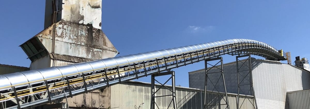 Galvanized RPS conveyor covers on a long elevated run curving over plant buildings, with a weathered silo behind