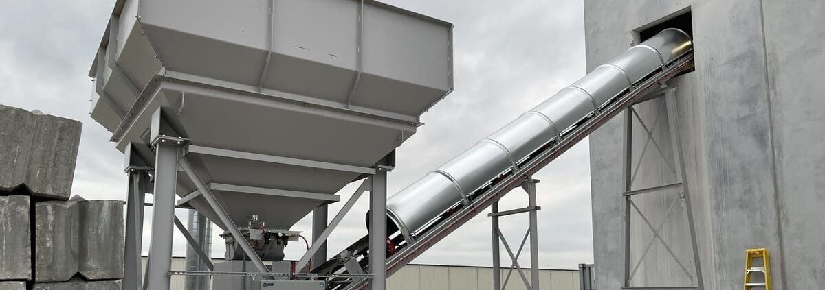 Inclined conveyor with smooth galvanized RPS covers rising from a batch hopper into a concrete plant building through a wall portal