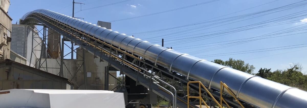 Smooth full-coverage galvanized RPS conveyor covers on a long inclined belt rising over an industrial plant, with maintenance walkway and access stairs alongside
